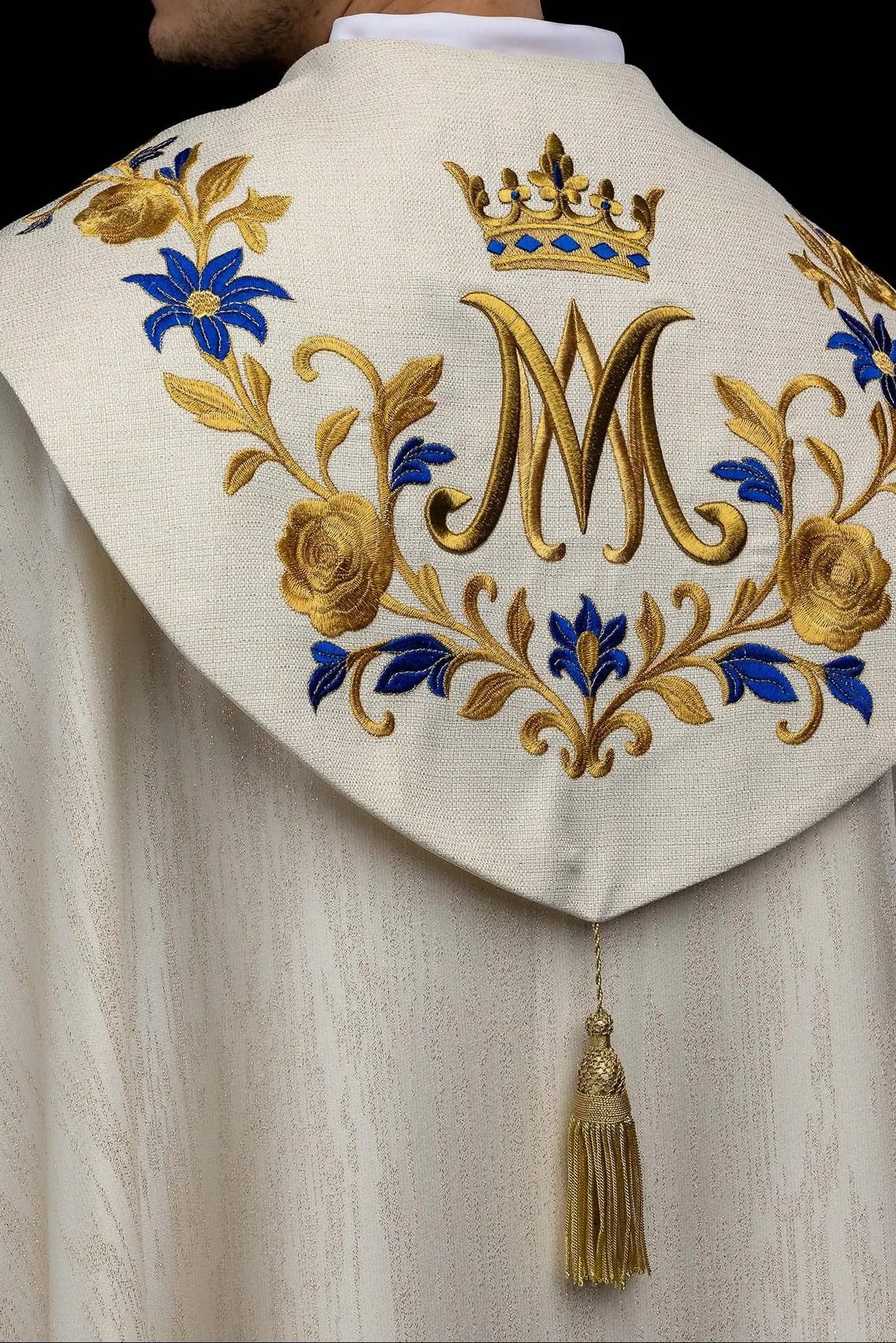 Ecru embroidered cope with Marian symbol, featuring gold and blue floral embroidery, a crowned 'M' monogram, and an ornate gold clasp with chain.